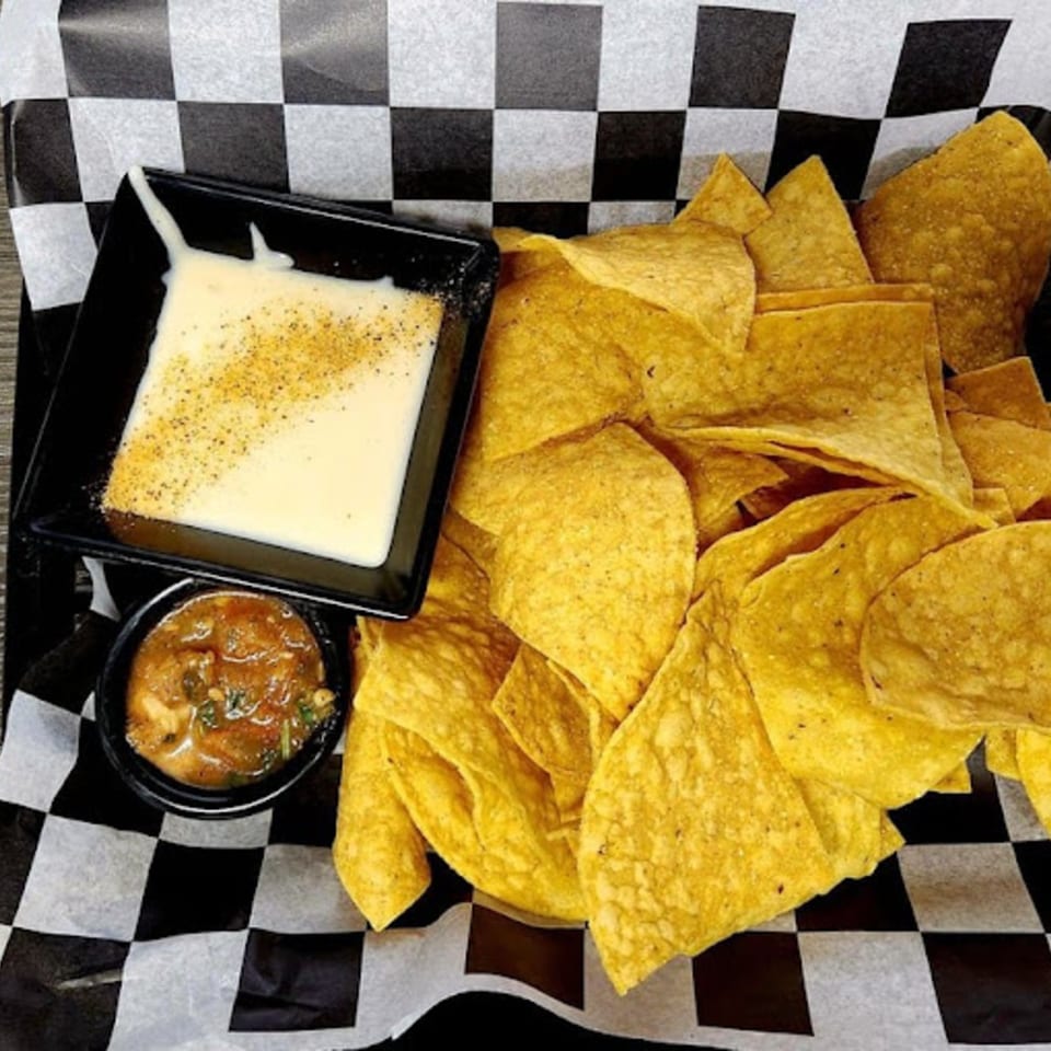 A basket of tortilla chips with a side of queso and salsa on checkered paper.