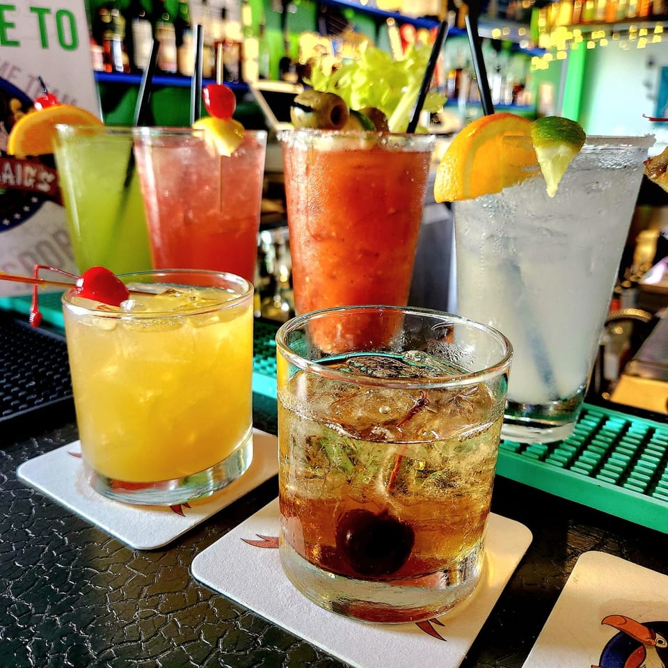 Five colorful cocktails, including a Bloody Mary, on a bar with bottles in the background.
