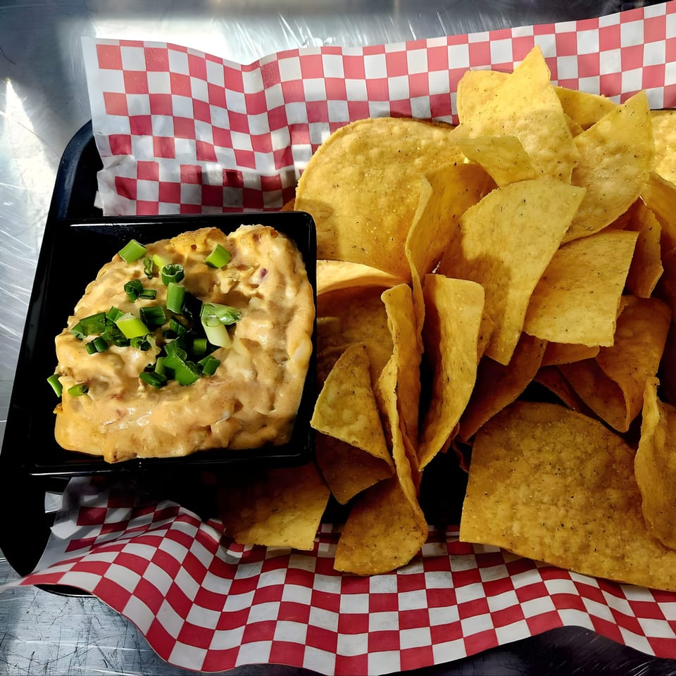 A black tray holds a bowl of cheesy dip topped with green onions and a pile of tortilla chips.